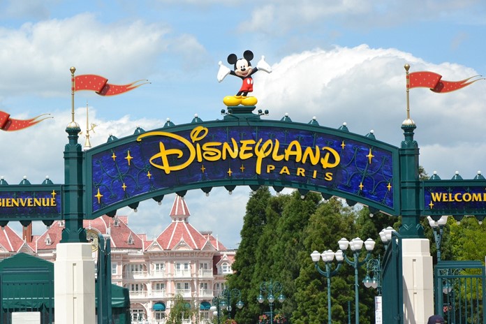 Das blaue Eingangsschild mit dem gelben Disneyland Paris Schriftzug. Micky Maus steht als Figur oben drauf. Das Disneyland Hotel ist im Hintergrund zu sehen.Bild